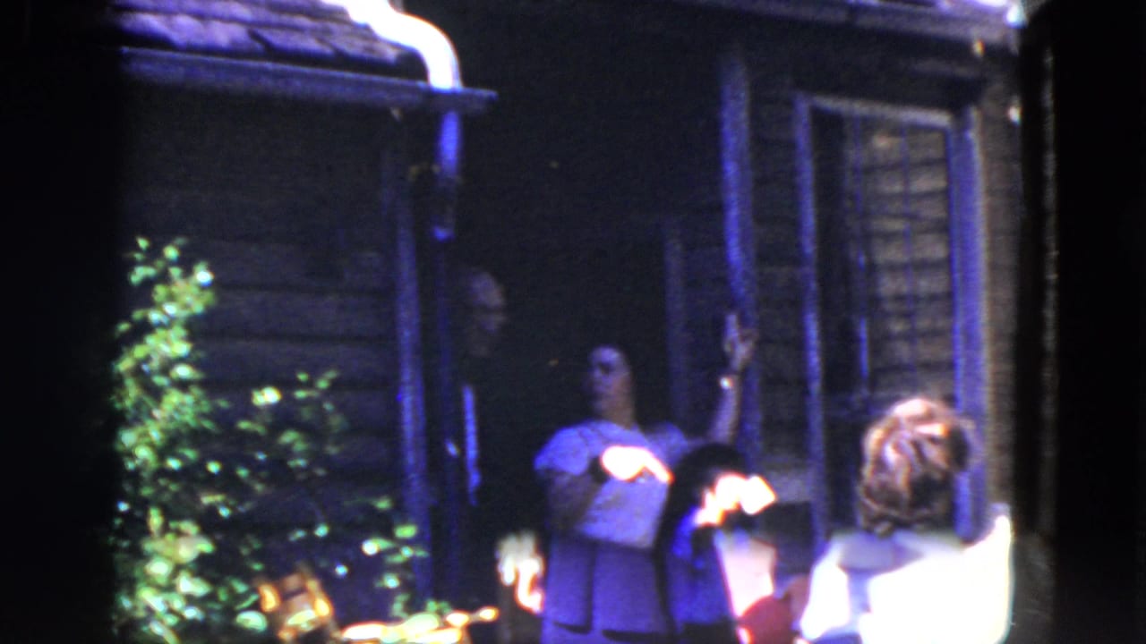 1964 8mm frame from Niagara Falls showing adults outside a wooden house, with a seated figure, plants, and strong shadow across the porch.
