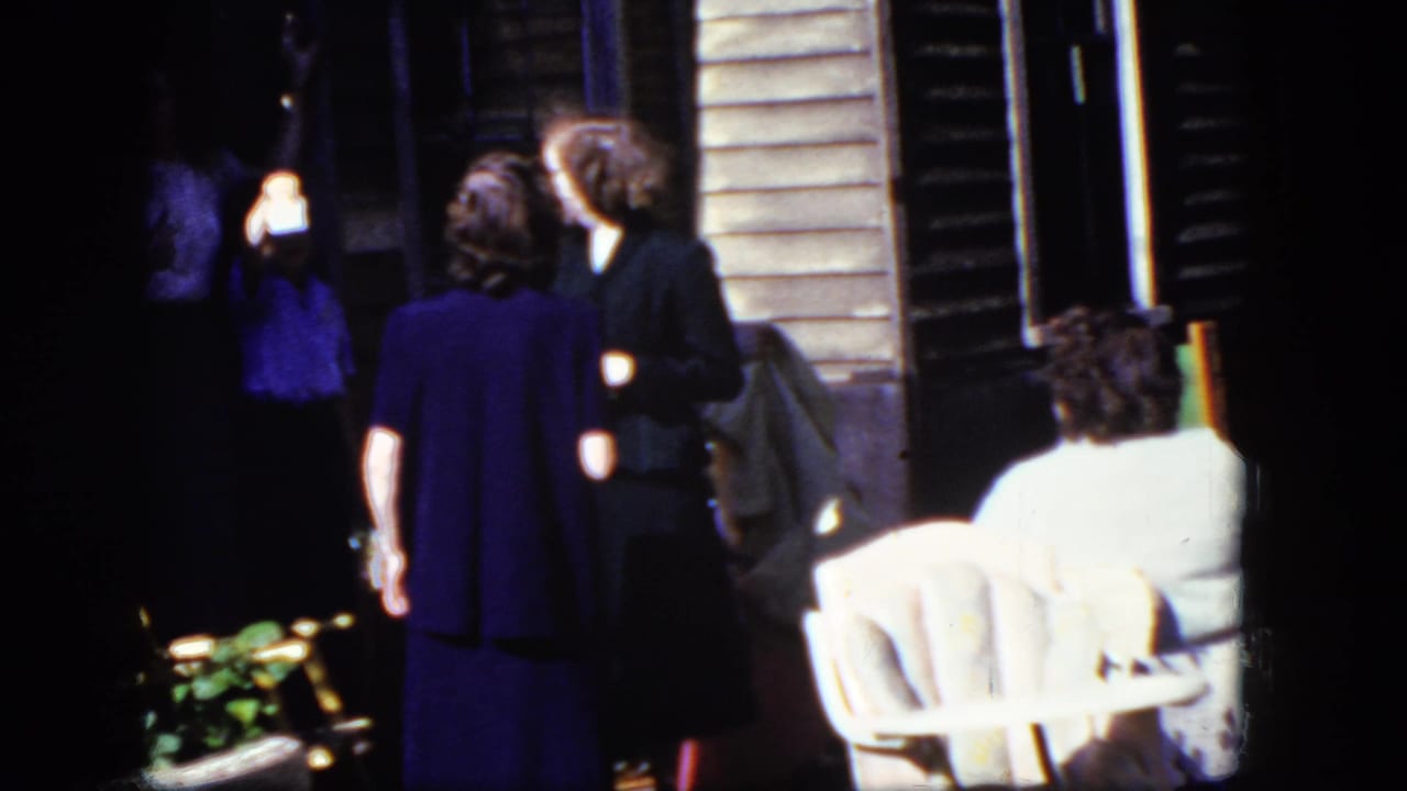 1964 Niagara Falls home-movie frame showing adults conversing near a porch, with a white outdoor chair and wooden siding visible.