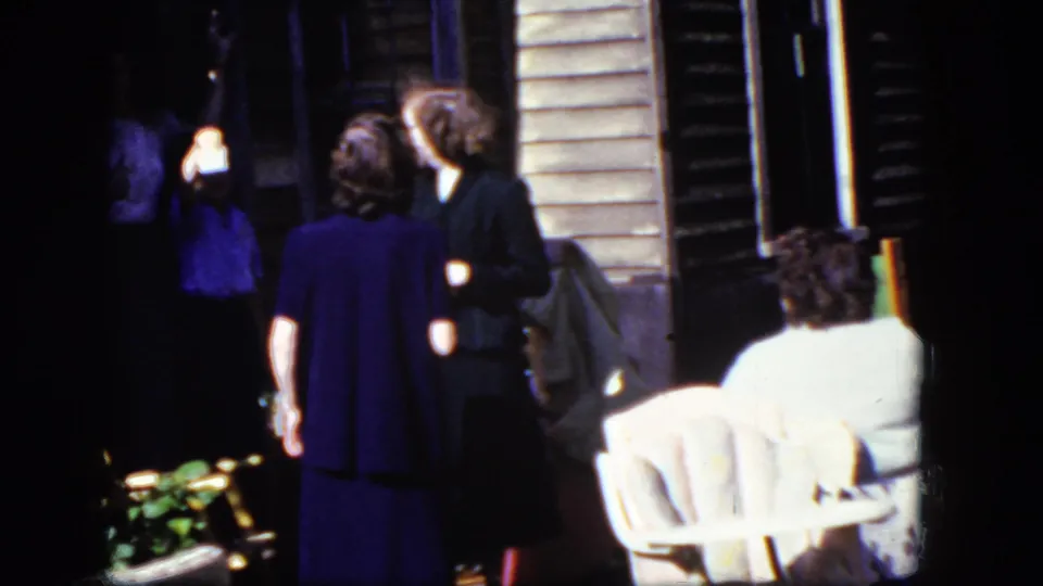 1964 Niagara Falls home-movie frame showing adults conversing near a porch, with a white outdoor chair and wooden siding visible.