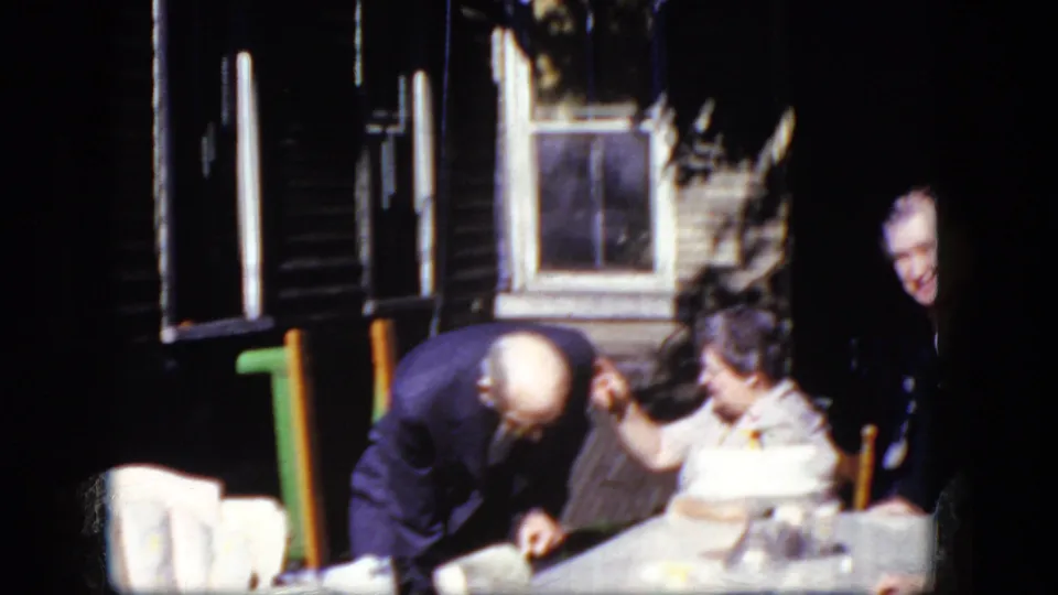 1964 Niagara Falls frame showing an older man in dark formal clothing leaning toward a seated woman at an outdoor table by a wooden house.