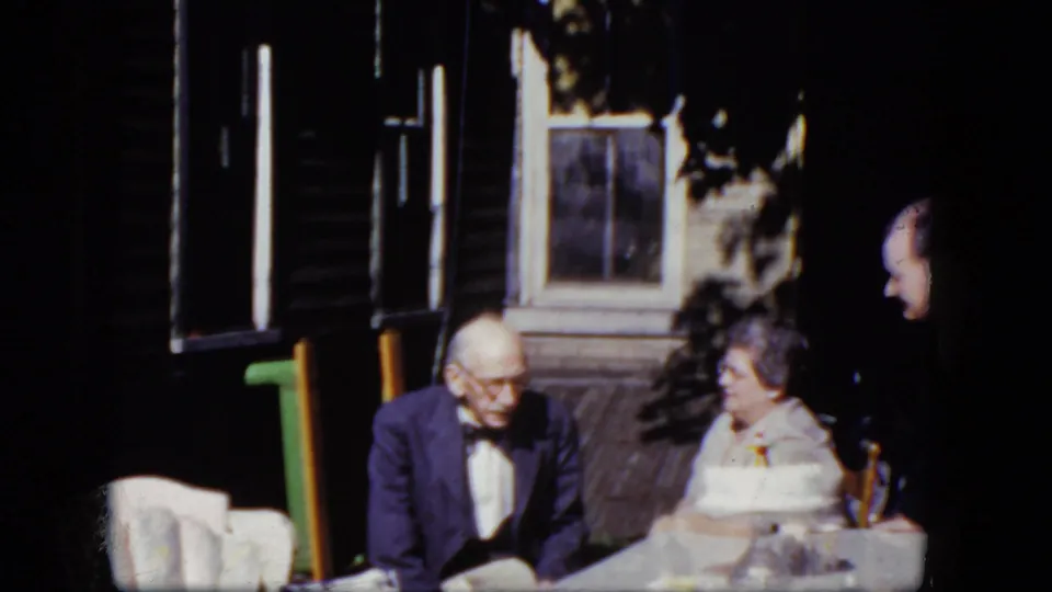 1964 Niagara Falls home-movie frame showing an older man in a bow tie seated beside a woman at a table outside a wooden house.