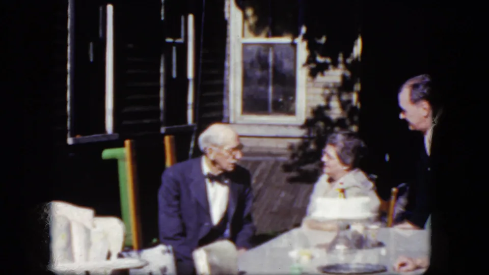 1964 8mm frame from Niagara Falls, New York, showing an older couple and a younger man conversing at an outdoor table on a porch.