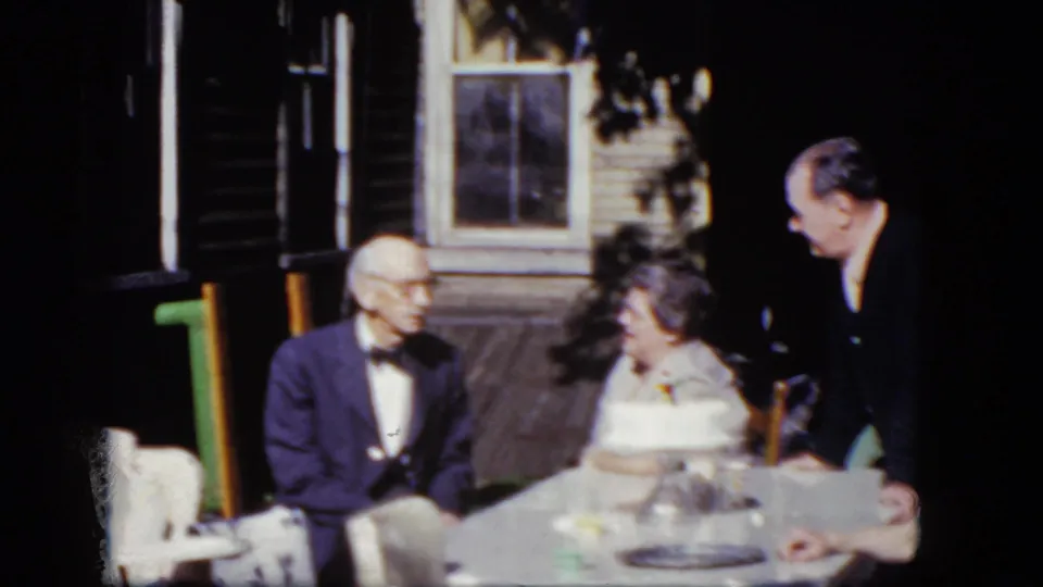 1964 Niagara Falls home-movie frame with an older couple seated at a table while a younger man in a dark suit leans in nearby.