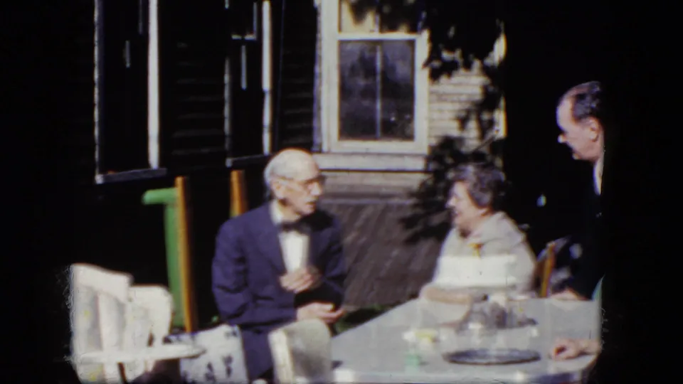 1964 8mm home-movie frame showing an older man gesturing at a porch table beside an older woman, with a younger man nearby.
