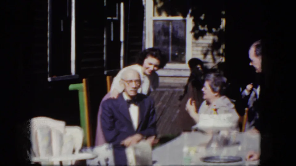 1964 Niagara Falls frame showing an older couple seated at a porch table as younger adults appear nearby in the home-movie scene.