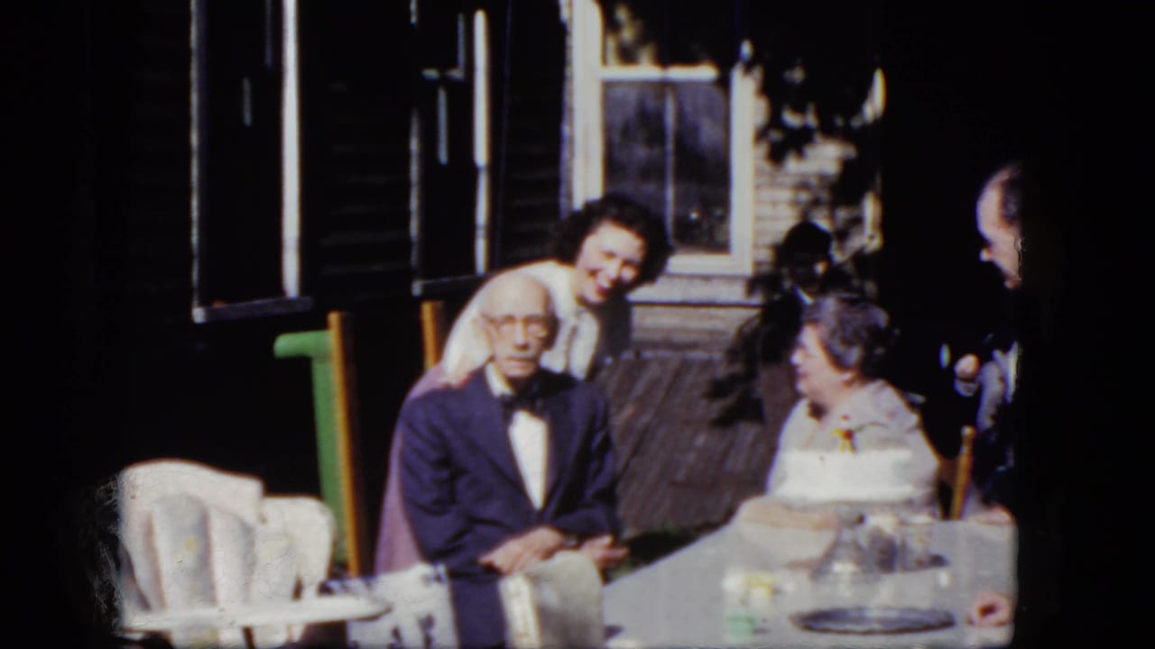 Elderly man in a suit sits at an outdoor table as women gather near him on a porch.