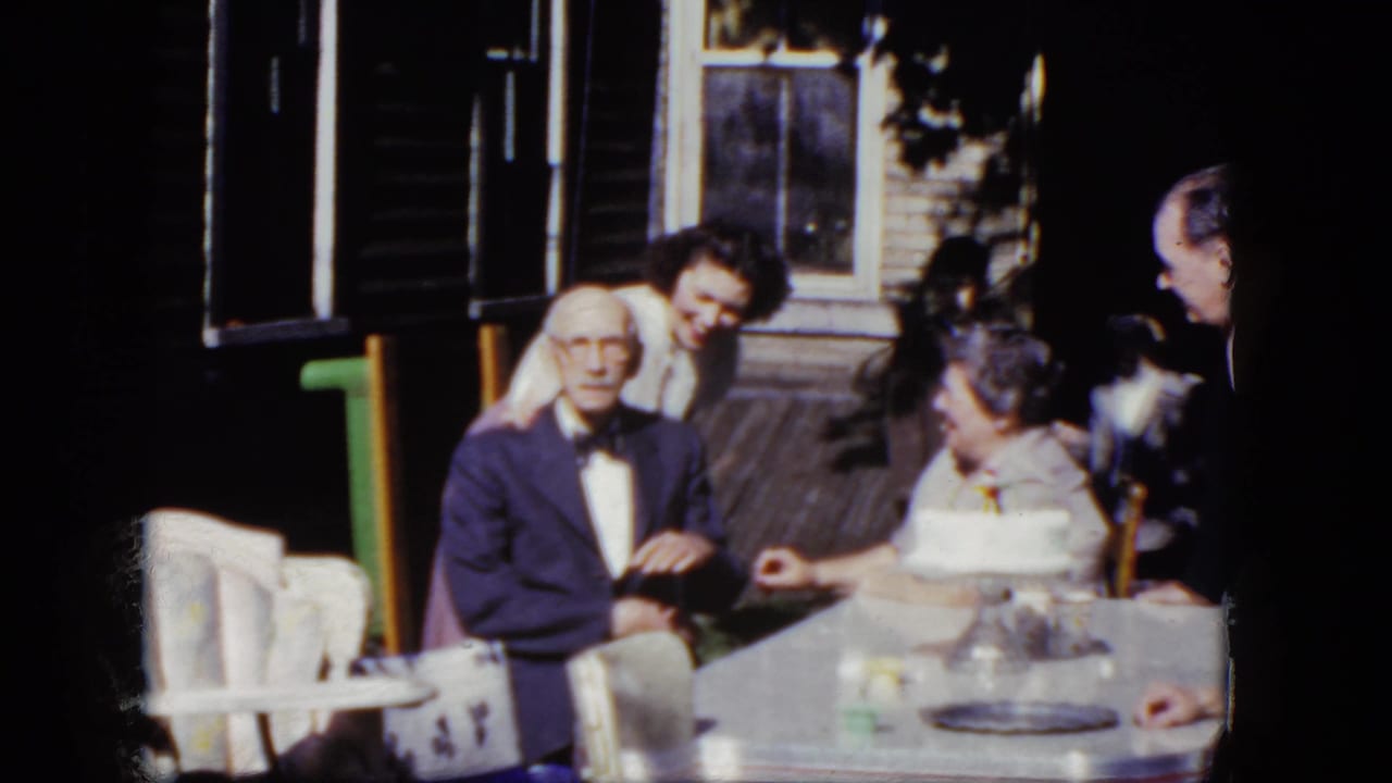 1964 Niagara Falls home-movie frame with an elderly man in a suit beside a birthday cake on a sunlit porch table.