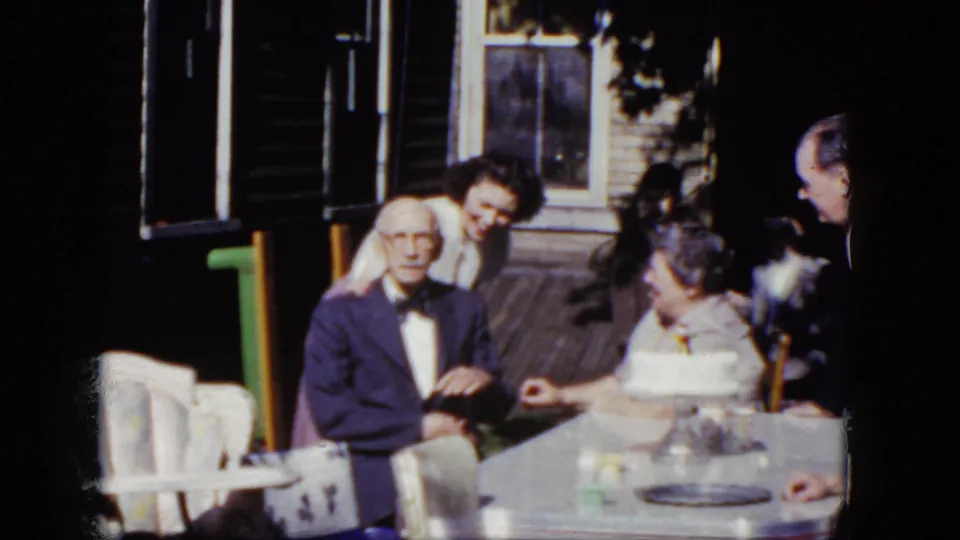 1964 Niagara Falls home-movie frame with an elderly man in a suit beside a birthday cake on a sunlit porch table.