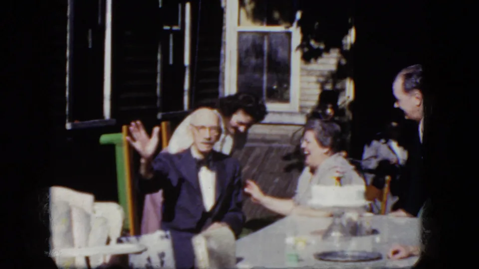 1964 8mm frame from Niagara Falls, New York, showing an elderly man waving at the camera during a porch birthday celebration.