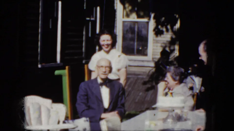 1964 Niagara Falls home-movie frame of a smiling woman behind an elderly man at a porch birthday table with cake visible.