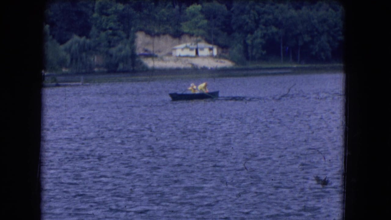 Two people in a small rowboat cross a rippled lake with a wooded shore behind them.