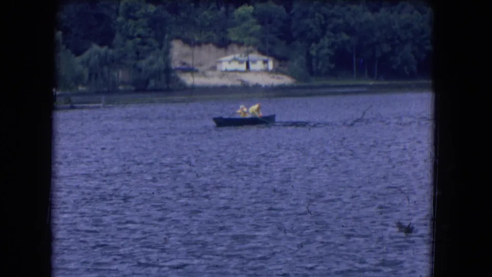 1964 8mm home-movie frame near Niagara Falls, New York, showing two people in yellow tops rowing across a lake below a wooded shoreline.
