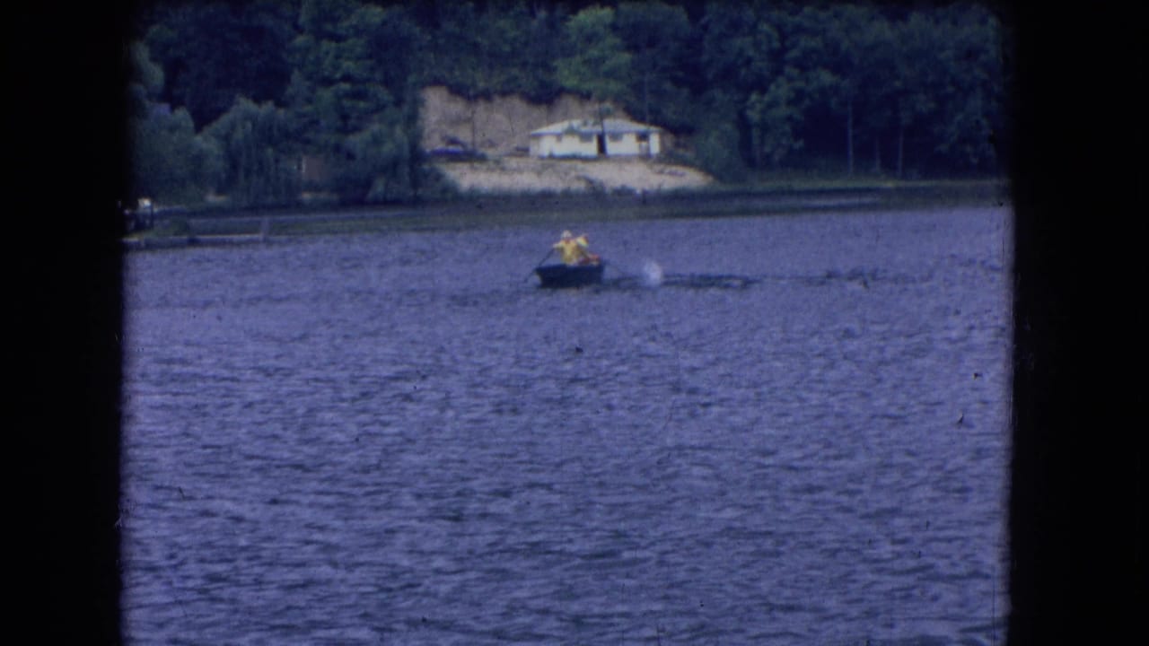 1964 8mm frame from Niagara Falls, New York, with two distant boaters on a lake and a pale house set back on the wooded bank.