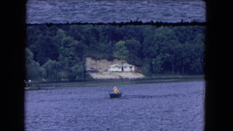 Home-movie frame from the 1964 Niagara Falls, New York clip, showing the rowboat smaller in the lake with trees and shoreline behind it.