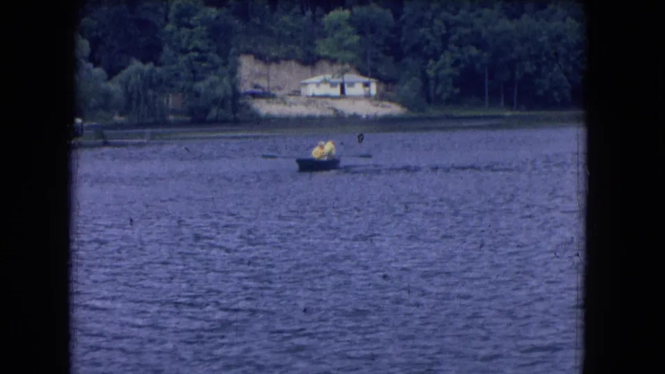 1964 8mm Niagara Falls, New York frame showing two distant people in a small rowboat on a lake with wooded shore and a house beyond.
