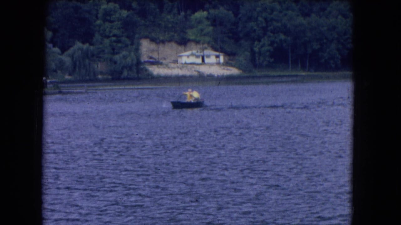 1964 8mm frame from Niagara Falls, New York, showing two distant rowers on a lake below a wooded shoreline and pale house.