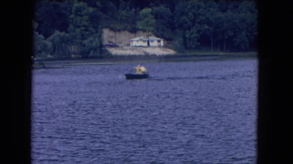 1964 8mm frame from Niagara Falls, New York, showing two distant rowers on a lake below a wooded shoreline and pale house.