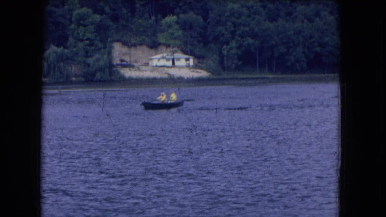 A small boat with two people moves across open water near a tree-lined shore and house.