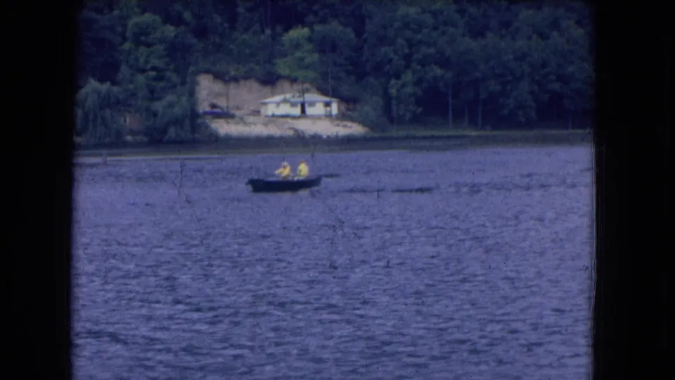 Distant 1964 home-movie view of two people rowing on a lake in Niagara Falls, New York, with shoreline trees and a house behind them.
