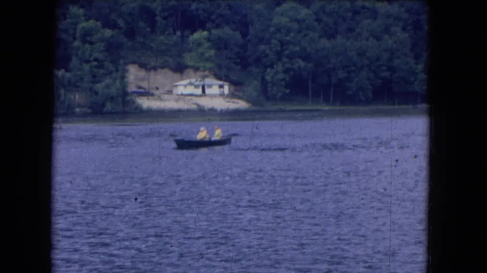 1964 8mm lake scene from Niagara Falls, New York, with two rowers in bright shirts passing a wooded bank and distant house.