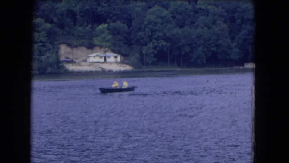 Final 1964 8mm frame of two people rowing across a Niagara Falls, New York lake, with a wooded shoreline and pale house in the distance.