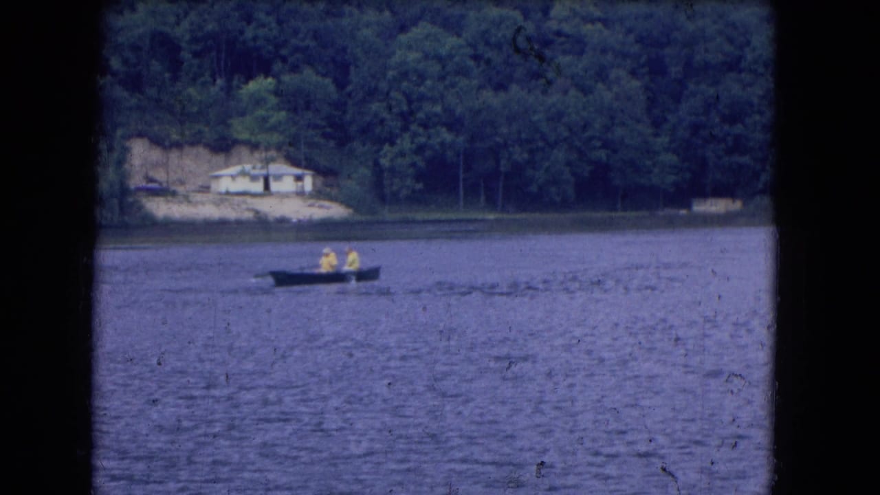 Two yellow-clad people in a small dark boat on a lake near a wooded shoreline.