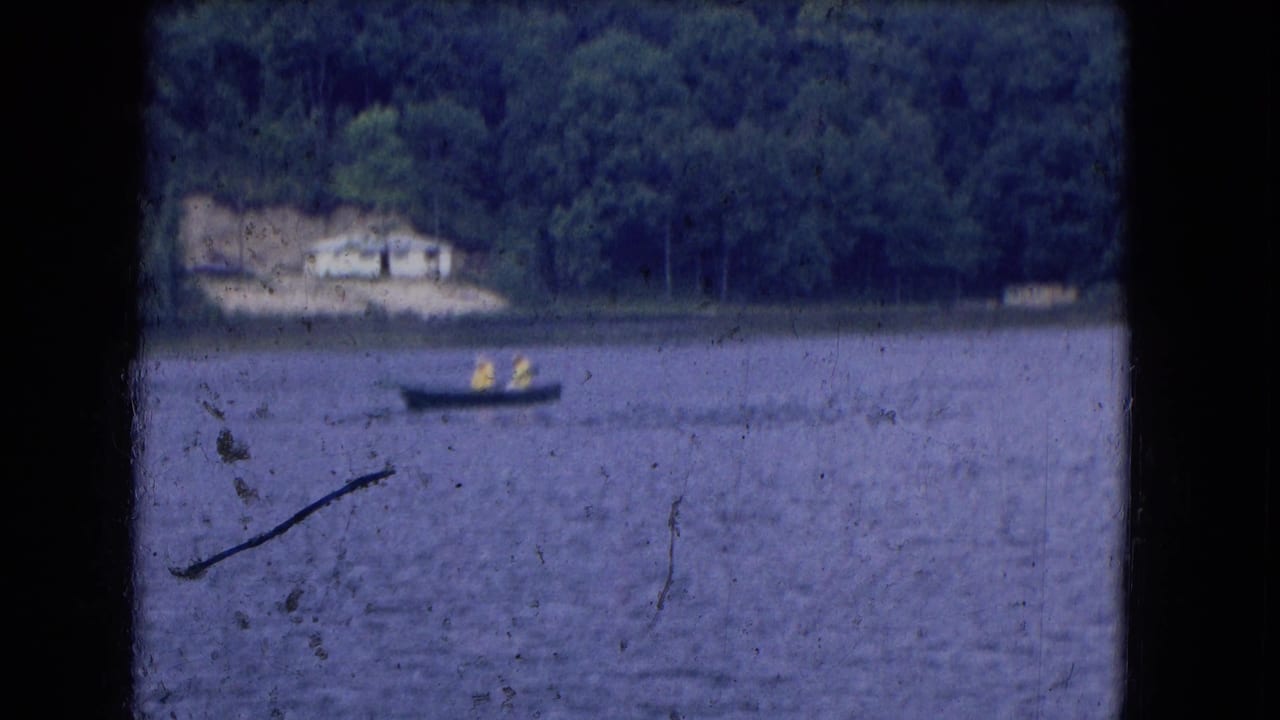 Distant 1964 8mm view of two people in yellow rainwear in a small boat on a lake, with a wooded shoreline and pale building behind.