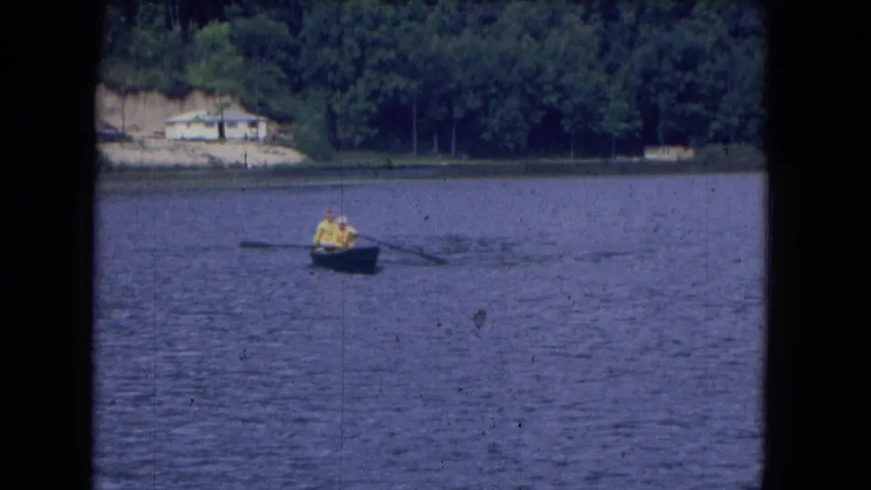 1964 home-movie frame showing two yellow-clad people rowing a small dark boat on a lake, viewed from shore at a distance.