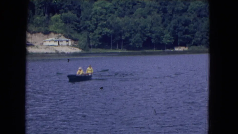 End frame of a 1964 8mm lake scene with two people in yellow rainwear rowing a small boat near a wooded shoreline.