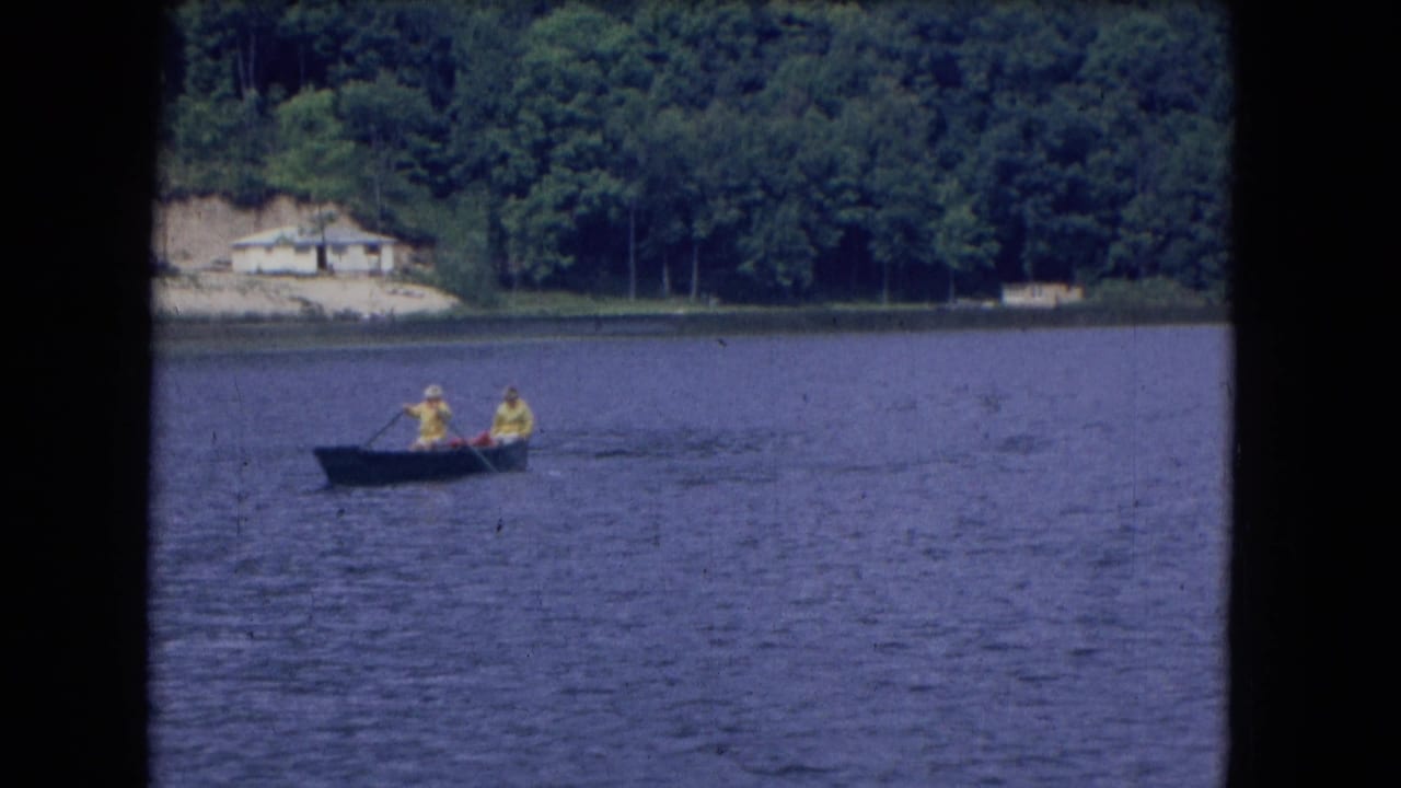 Two people in a small rowboat on a calm lake near a wooded shoreline.
