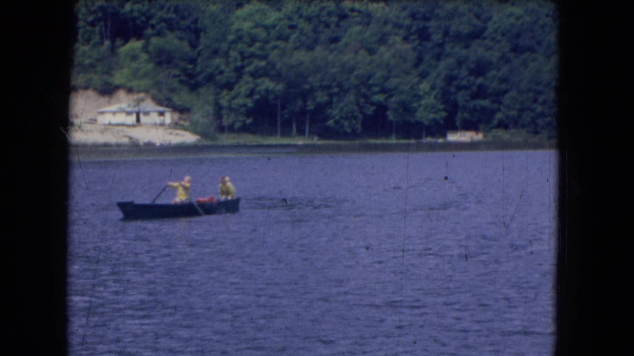 Two people in yellow tops row a dark boat across a lake in this 1964 8mm frame from the credited Niagra Falls, New York footage.
