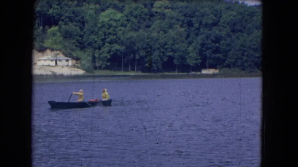 A distant 1964 8mm view shows two people rowing a small boat across calm lake water near a wooded shoreline.