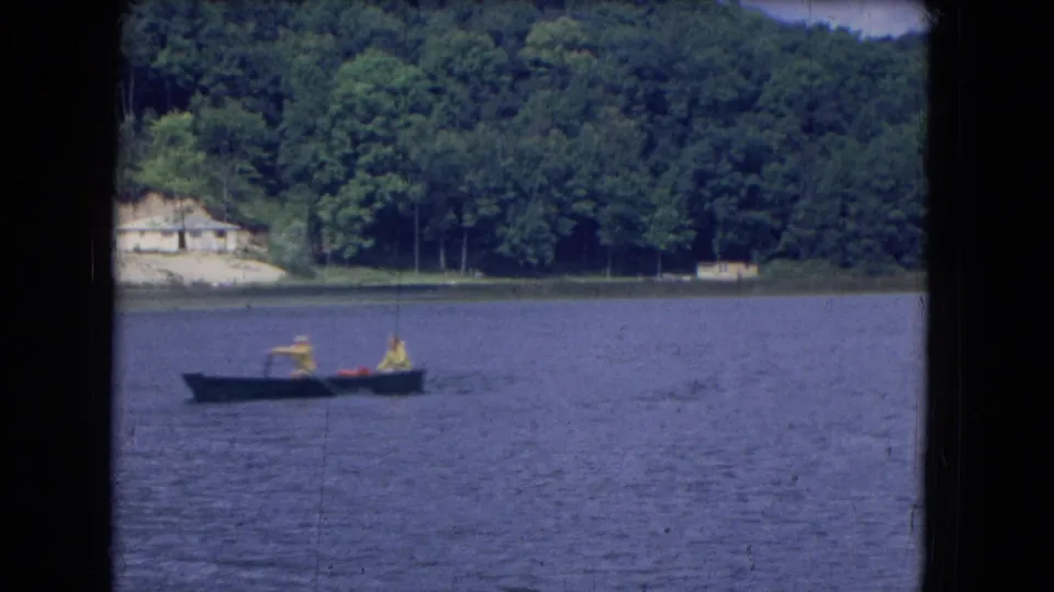 The 1964 home-movie frame shows two people in a small rowboat on a lake, with trees and a pale shoreline building behind them.