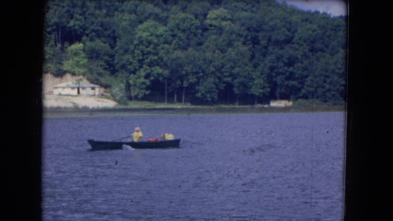 Small boat on a lake with distant people, wooded shore, and a pale building on the hillside.