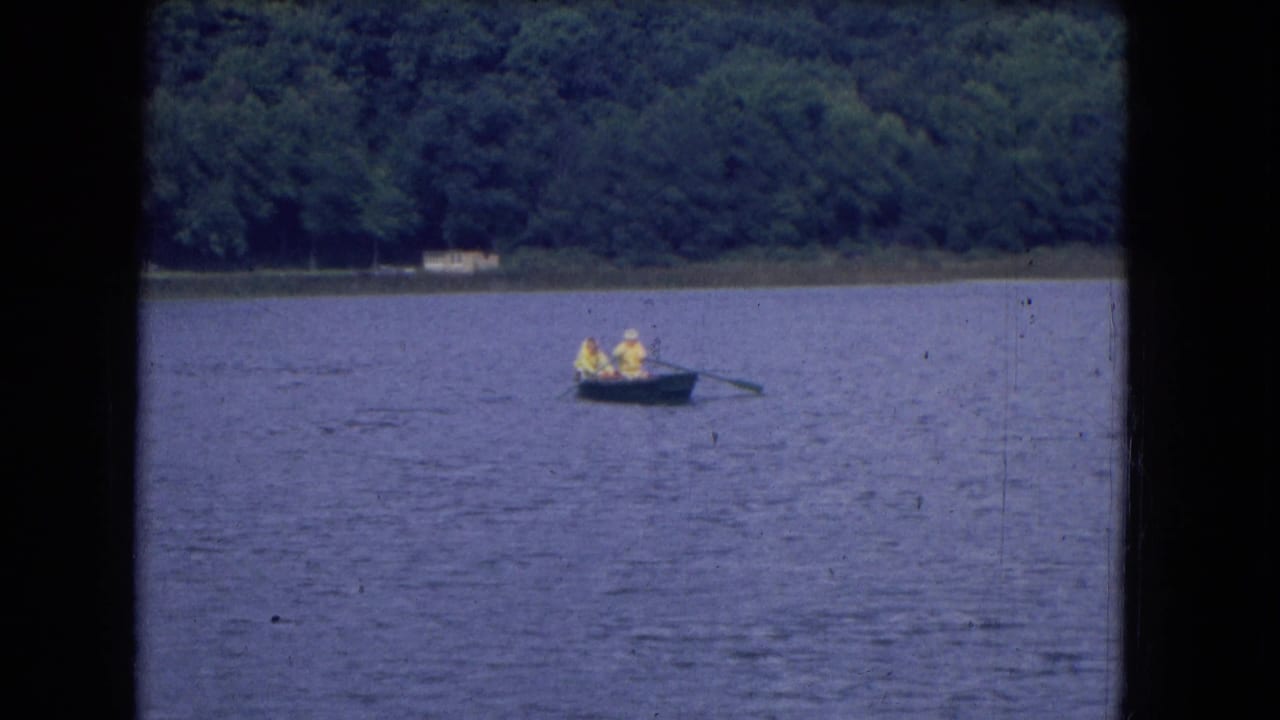 Distant rowboat with people in yellow on open lake water near a tree-lined shore.