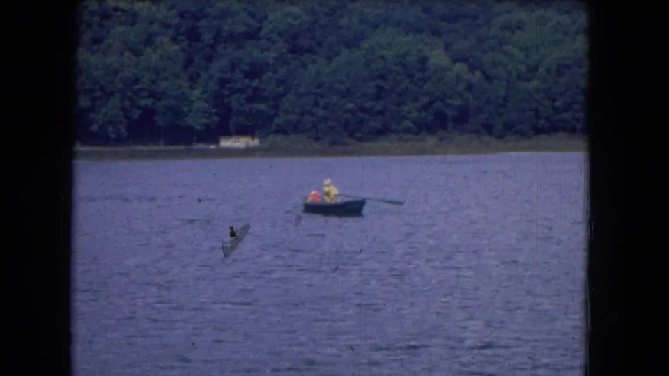 1964 8mm frame of distant boaters rowing on a lake near Niagra Falls, with trees along the shore and visible film marks.