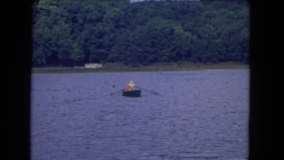 1964 home-movie still from Niagra Falls, New York, showing distant people in a small boat rowing across a calm wooded lake.