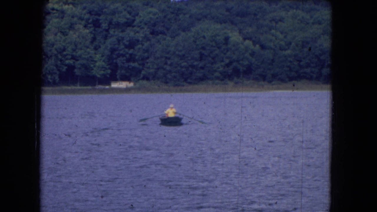 A distant person rows a small boat across a lake with trees on the far shore.