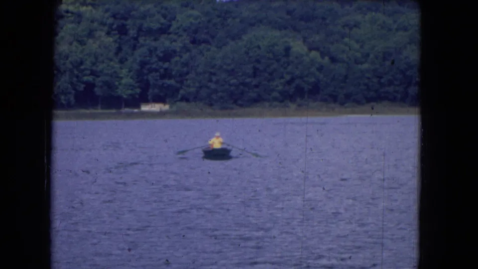1964 8mm home-movie frame from Niagara Falls, New York, showing a distant person in yellow rowing on a calm lake.