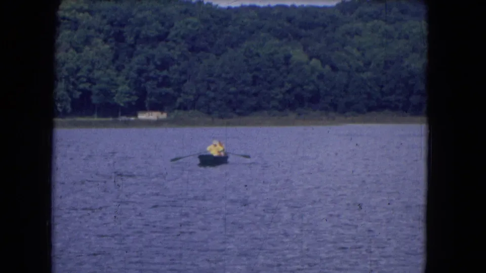 1960s 8mm frame showing a person in yellow rowing a small boat across lake water with wooded shoreline in the background.