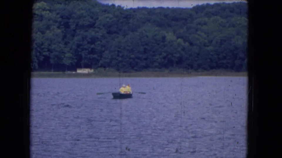 Final 1964 home-movie frame of the distant rower on a lake, with oars visible and trees lining the far shore.