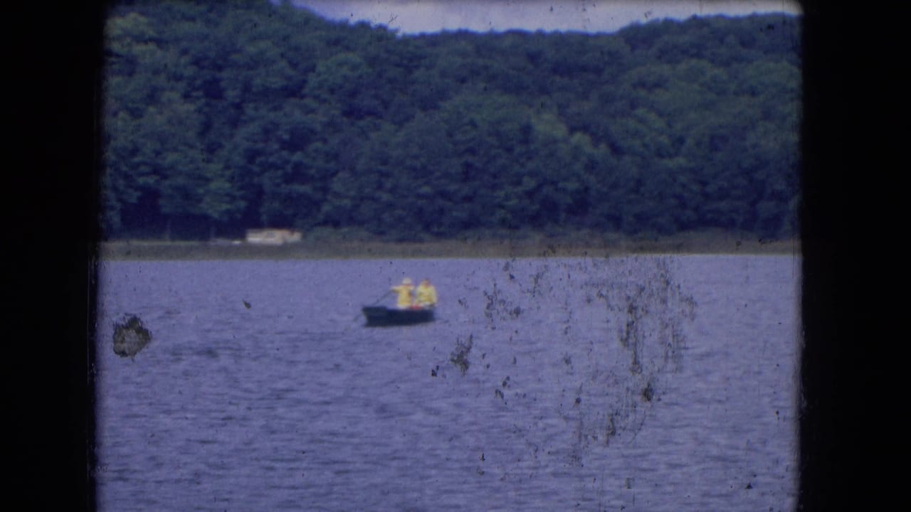 1964 8mm home-movie frame showing a small boat with two distant people on calm water near a forested shoreline.