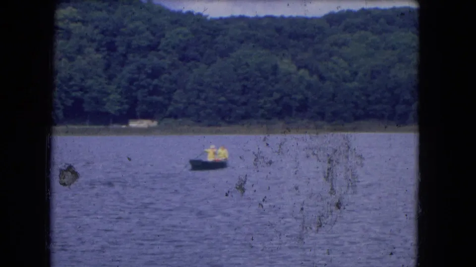 1964 8mm home-movie frame showing a small boat with two distant people on calm water near a forested shoreline.