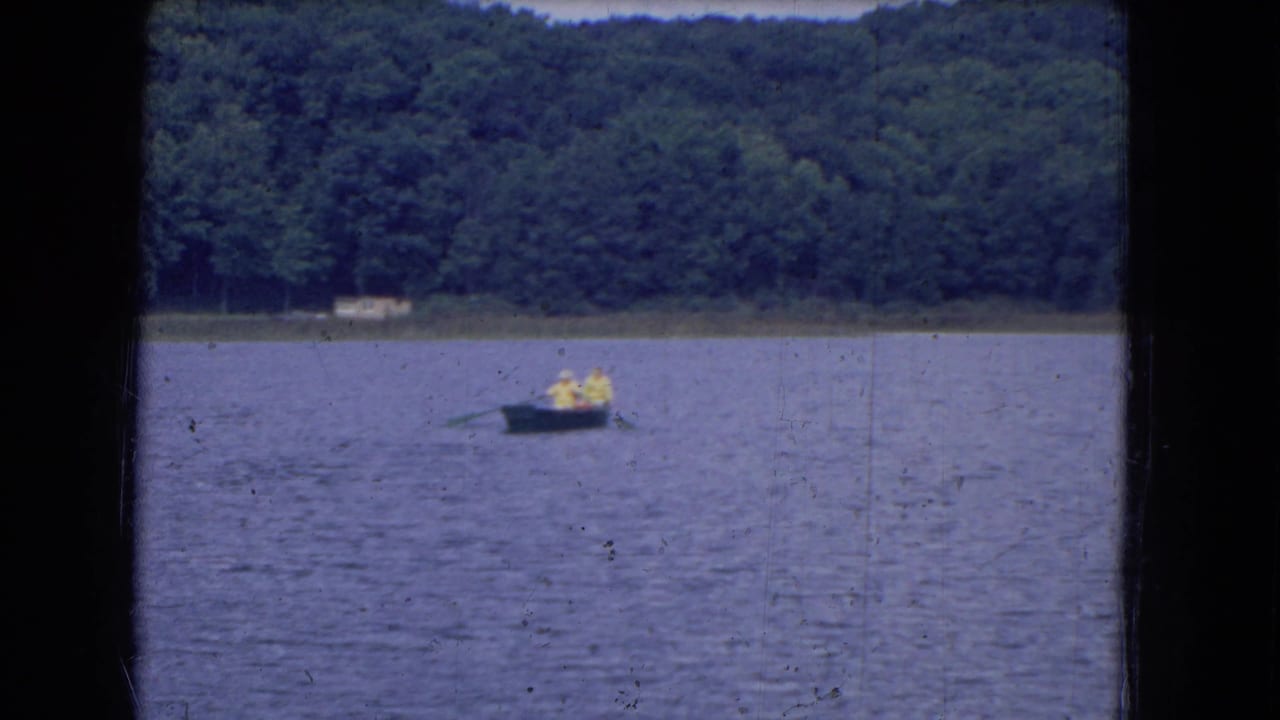 1964 8mm frame of a small boat carrying two distant people across a lake, backed by dense trees along the shore.