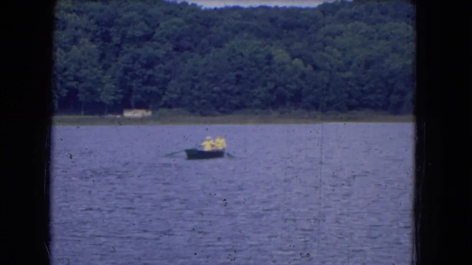 1964 8mm frame of a small boat carrying two distant people across a lake, backed by dense trees along the shore.