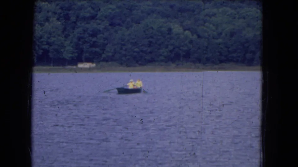 Distant 1964 lake boating view with two people in a small craft, framed by water, trees, and 8mm film wear.