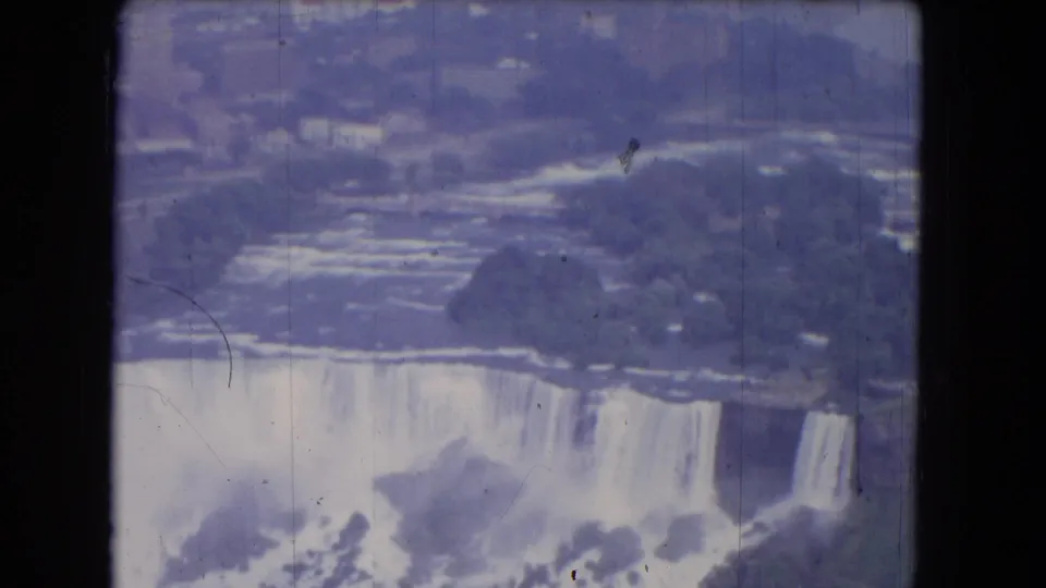 1964 Niagara Falls frame showing the cascade from above, with mist, trees, shoreline, and visible film wear.