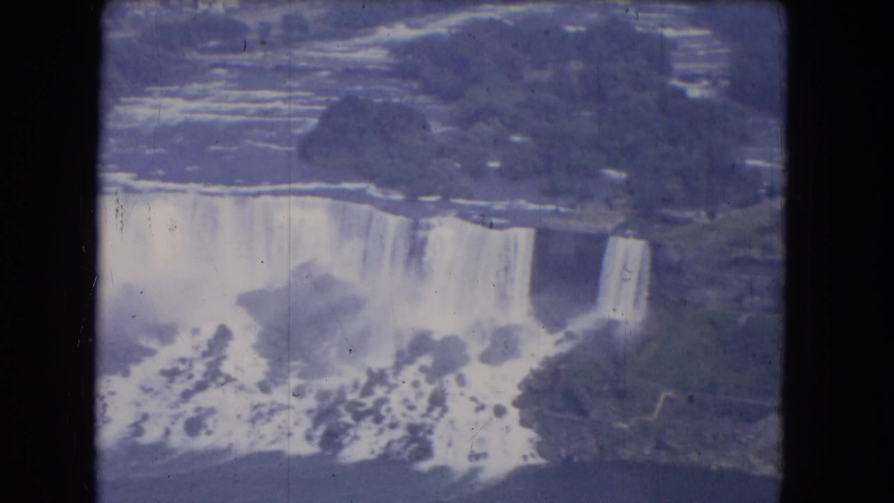 Aerial view of broad waterfalls dropping into mist with rocky shoreline nearby.