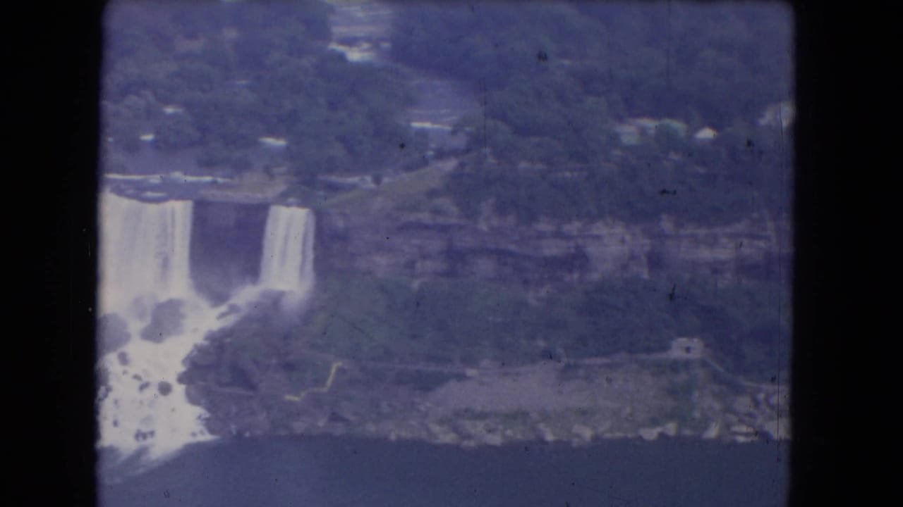 1964 Niagara Falls aerial frame showing falling water, shoreline paths, mist, and soft blue-purple 8mm color.