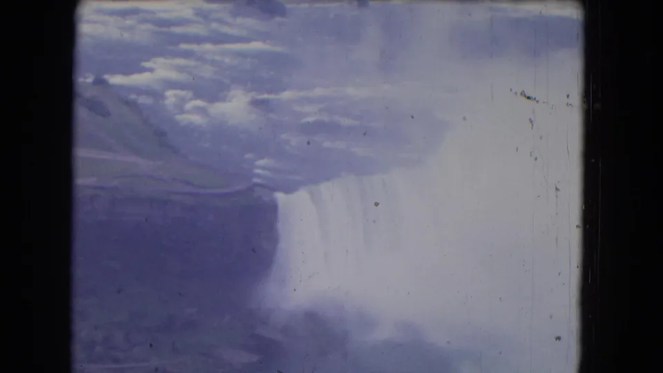 1964 8mm aerial view over Niagara Falls with dense white mist, dark water, and faded blue-purple film tones.